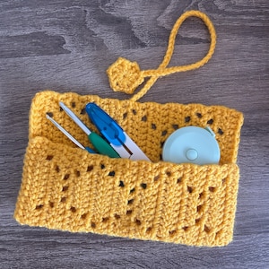 May include: A handmade, mustard yellow crochet organizer with a decorative openwork design. The organizer holds crochet hooks, scissors, and a light blue measuring tape. A loop and flower-shaped embellishment are attached to the top.