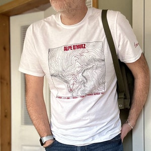 May include: White t-shirt featuring a black and white topographic map design of the Alpe d'Huez mountain pass. The text "ALPE D'HUEZ" is in red, and the text "13.8 KM / L'ALPE / 1860m / TOUR DE FRANCE DEPUIS 1952" is in black.