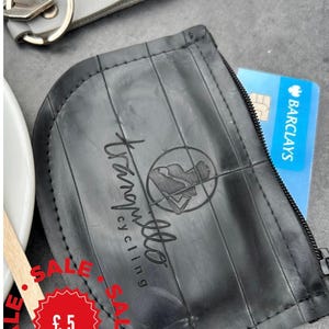May include: A black, semi-circular zippered pouch crafted from recycled rubber, featuring the text 'tranquillo cycling' and a circular logo. A blue Barclays bank card is partially inserted into the zipper opening of the pouch.