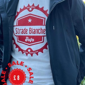 May include: A white t-shirt featuring a red circular graphic with a gear-like border, the text Strade Bianche, Eolo, Dal 2007, and Siena, Toscana. The shirt is worn under a dark navy blue hooded jacket. A red circular sale badge displays the text Sale and £ 8.