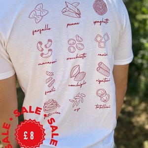 May include: A white short-sleeved t-shirt featuring a red graphic print on the back. The design displays various pasta shapes including farfalle, penne, spaghetti, macaroni, orecchiette, ravioli, fusili, conchiglie, rigatoni, fettuccine, orzo, and tortellini.