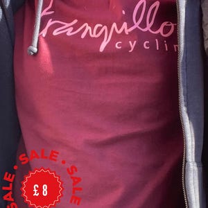 May include: A maroon t-shirt featuring the text "tranquillo cyclin" in a light pink script font. The shirt is worn under a dark navy blue zip-up hoodie. A red circular graphic with a jagged edge displays the text "SALE" and the price "£ 8".
