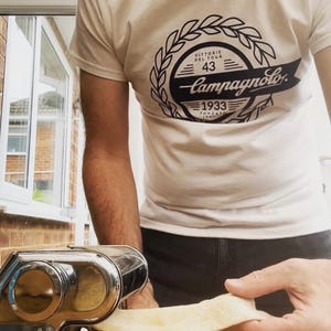 May include: A person wearing a white t-shirt with a black and white logo that says "Campagnolo" and "VITTORIE DEL TOUR 43 1933 FONDATO ITALIANO". The person is using a silver pasta maker to roll out a sheet of dough.