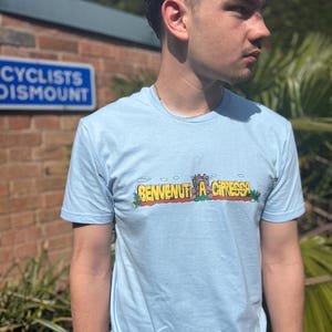 May include: A light blue t-shirt with a cartoon graphic of a castle and the text "BENVENUTI A CIPRESSA".