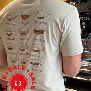 May include: A beige short-sleeved t-shirt featuring a brown graphic print on the back. The design displays a grid of twelve coffee cup illustrations, each labelled with its specific name, including Espresso, Doppio, Ristretto, Macchiato, Americano, Long Black, Flat White, Cappuccino, Cafe Latte, and Latte Macchiato.
