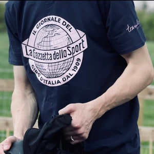 May include: A navy blue t-shirt with a pink and white logo featuring a globe and the text "Il Giornale Del La Gazzetta dello Sport Giro d'Italia Dal 1909".