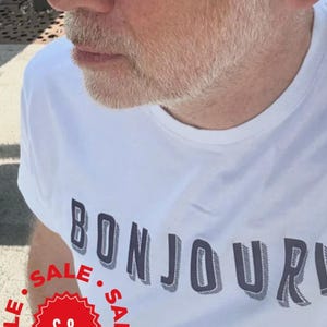 May include: A white short-sleeved t-shirt featuring the word BONJOUR in dark blue block letters with a shadow effect. The shirt is worn by an individual with a light beard wearing black-framed sunglasses with dark lenses and a red sale sticker.