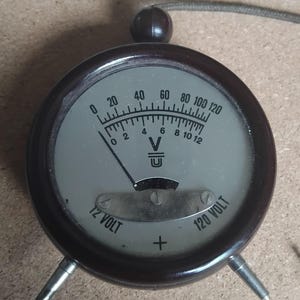 May include: A vintage voltmeter with a dark brown casing and a light grey face. The gauge displays voltage readings from 0 to 120 volts, with a needle indicating the current level. It has two metal prongs and a small knob.