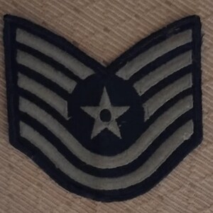 May include: A dark blue military patch featuring a star and five light grey stripes. The patch has a curved top and a pointed bottom. The star has a small circle in the centre.