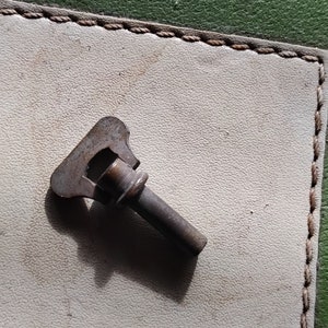 Puede incluir: Una pequeña llave de metal oxidada con una cabeza curva única. La llave está sobre una superficie blanca con un borde cosido marrón.