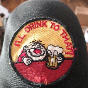 Puede incluir: Una gorra de béisbol negra con un parche bordado en rojo, amarillo y blanco que dice "Brindo por eso". El parche presenta un personaje de dibujos animados sosteniendo un vaso de cerveza.