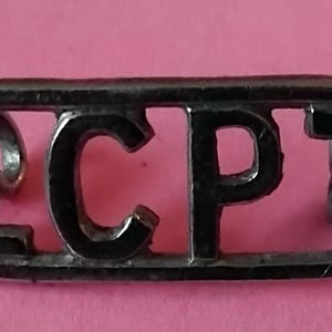May include: A silver-coloured metal pin featuring the letters "LCPT" in a cut-out design. The pin is rectangular with rounded edges and is set against a pink background. The letters are in a bold, sans-serif font.
