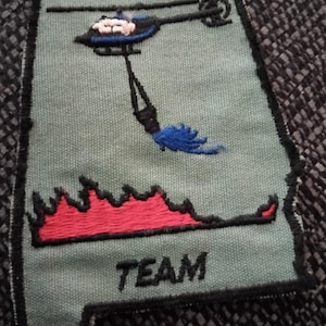Può includere: Una toppa ricamata verde con il testo "Helitack Team" e un elicottero blu che lascia cadere un secchio d'acqua su una fiamma rossa.
