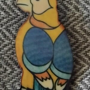 May include: A ceramic bird brooch in yellow, blue and orange. The bird is facing left and has a small, round eye. The bird's wings are spread out and its tail feathers are visible.