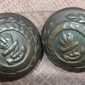 May include: Two antique metal buttons with a circular design. Each button features a stylized anchor with a crown and a wreath of leaves around the edge.