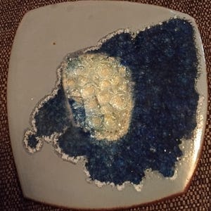 May include: A square ceramic plate with a blue and white design. The design features a large, irregular shape with a white centre and a blue border. The centre of the shape has a textured, bubbly appearance.