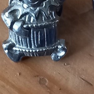 Può includere: Una statuetta in metallo argentato di una persona con un cappello che tiene una botte. La statuetta è in piedi su una superficie di legno.