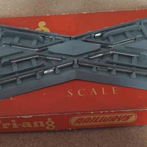 May include: A grey plastic model railway track crossing, placed on a red box. The box is printed with the words "SCALE" and "Tri-ang Railways". The track piece is designed to allow model trains to cross each other.
