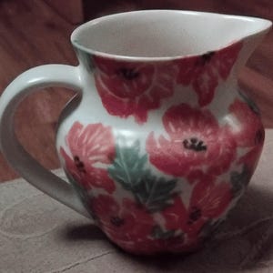 May include: A white ceramic jug with a curved handle and a spout. The jug is decorated with red poppy flowers and green leaves. The interior is white.