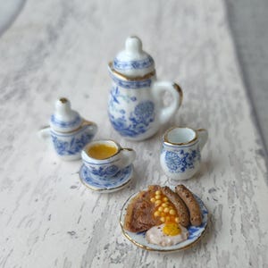 May include: A miniature porcelain tea set with blue floral patterns and gold edging. The set includes a teapot, creamer, sugar bowl, teacup and saucer, and a plate of miniature breakfast food. The plate has a fried egg, beans, and sausages.