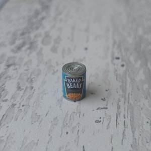 Peut inclure: Une petite boîte de conserve de haricots cuits. La boîte a un couvercle argenté et une étiquette bleue avec les mots "BAKED BEANS" en blanc. L'étiquette présente également une illustration de haricots cuits.