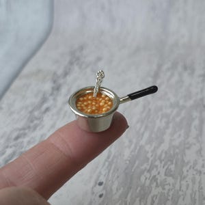 May include: A tiny silver saucepan filled with baked beans, with a small spoon inside. The handle is black. The beans are in a light orange sauce. The saucepan is resting on a finger.