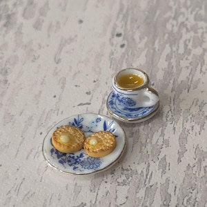 May include: Miniature porcelain tea set with two tarts. The white teacup and saucer have blue floral patterns and gold trim. The small plate holds two tarts with a yellow filling. A charming, detailed dollhouse accessory.