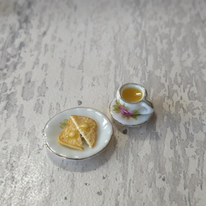 May include: A miniature ceramic set featuring a white plate with two slices of toasted bread topped with yellow butter, and a matching white teacup on a saucer decorated with a pink rose floral pattern and gold-toned trim on the edges.
