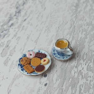 May include: A miniature teacup and saucer with a blue and white floral pattern, filled with a light brown liquid, and a plate with a variety of miniature biscuits, including a pink and white ring biscuit, a chocolate biscuit, and several round biscuits.