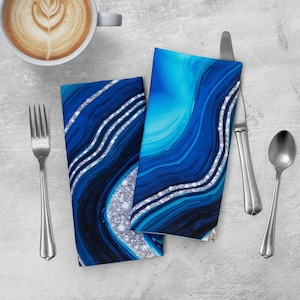 May include: Two blue and silver abstract patterned napkins with a white background. The napkins are folded and placed on a gray surface with a fork, knife, and spoon.
