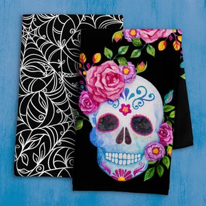 Puede incluir: Dos toallas de cocina negras, una con un diseño de telaraña blanca y la otra con un diseño de calavera de azúcar colorida y flores.