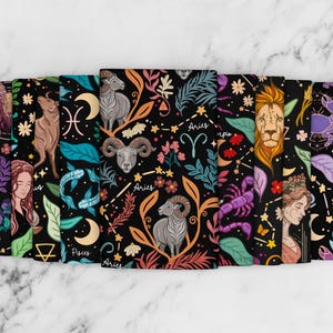 May include: A collection of notebooks with a black background and colorful zodiac illustrations. Each notebook features a different zodiac sign, including Aries, Taurus, and Leo, with floral and celestial designs. The notebooks are arranged on a white marble surface.