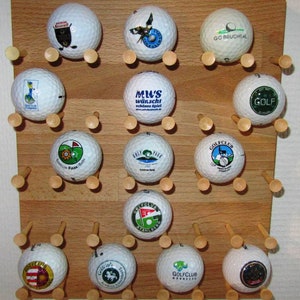Op de afbeelding: Een houten display met vijftien golfballen, elk met een ander logo of tekst, waaronder "GOLF" en "GOLFCLUB". De ballen worden op hun plaats gehouden door houten pinnen. De tekst "MWS wünscht schönes Spiel" is ook zichtbaar.