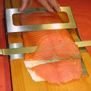 Könnte beinhalten: Ein Lachsschneider aus Edelstahl wird verwendet, um ein Lachsfilet auf einem Holzschneidebrett zu schneiden. Der Lachs hat eine leuchtend orange Farbe, und der Schneider wird von einer Hand gehalten. Das Schneidebrett ist aus hellem Holz.