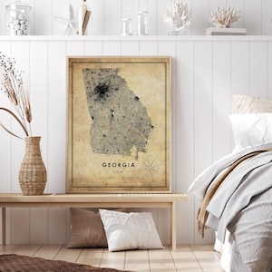 May include: A framed map of Georgia, USA, with a vintage aesthetic. The map is in shades of beige and black, with the state's outline and city details visible. The text "GEORGIA USA" and coordinates are at the bottom. The frame is a light brown color.