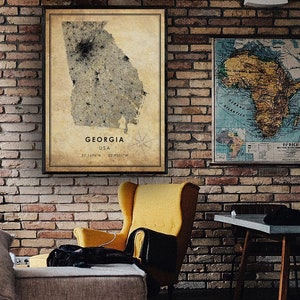 May include: A framed map of Georgia, USA, with a vintage aesthetic, hangs on a brick wall. The map features the state's outline and coordinates. A second framed map of Africa is also visible. A yellow armchair and a small table are in the foreground.