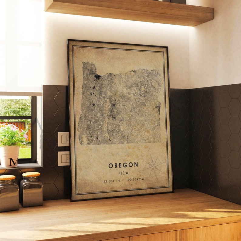 Puede incluir: Mapa enmarcado de estilo vintage de Oreg&oacute;n, EE. UU., con coordenadas y una rosa de los vientos. El mapa est&aacute; en un marco negro y descansa sobre una superficie de madera. El fondo incluye una ventana, frascos y una pared de azulejos oscuros.