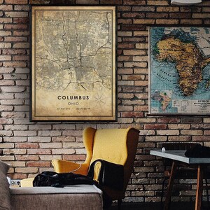 May include: A vintage-style map of Columbus, Ohio, with a compass rose and the coordinates 39.9412&deg;N, 82.9988&deg;W. The map is printed on aged paper and framed in a black frame. The map is hanging on a brick wall next to a yellow armchair and a wooden table.