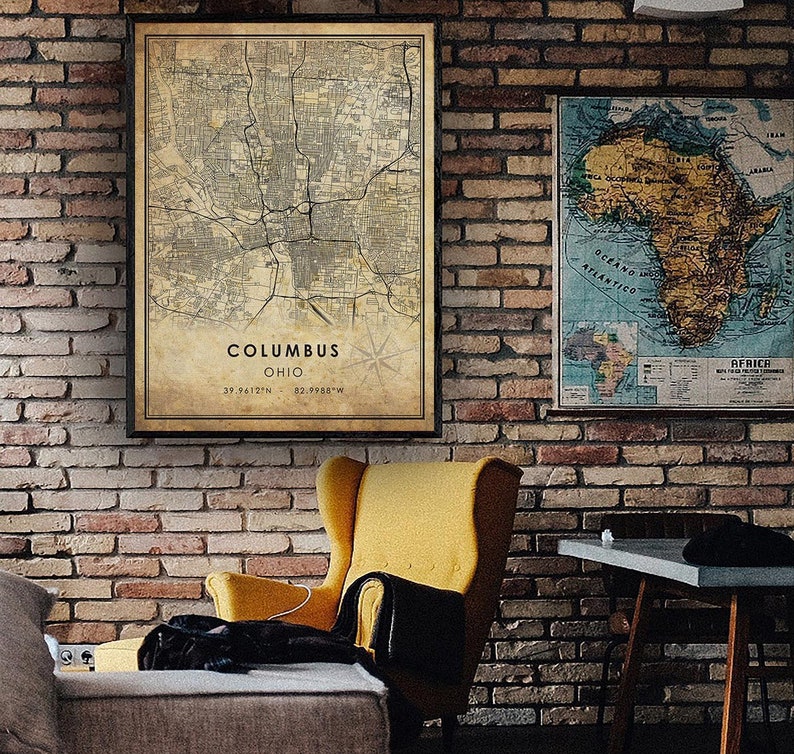 May include: A vintage-style map of Columbus, Ohio, with a compass rose and the coordinates 39.9412&deg;N, 82.9988&deg;W. The map is printed on aged paper and framed in a black frame. The map is hanging on a brick wall next to a yellow armchair and a wooden table.
