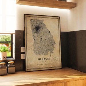 May include: Framed map print of Georgia, USA, in a neutral color palette. The map features a detailed depiction of the state's geography, with the name "Georgia" and coordinates displayed. The print is in a black frame and is displayed on a wooden surface.