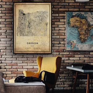 Puede incluir: Un mapa enmarcado de Oreg&oacute;n, EE. UU., con coordenadas y una rosa de los vientos, cuelga de una pared de ladrillo. El mapa tiene una est&eacute;tica vintage con un fondo beige y un marco marr&oacute;n oscuro. Tambi&eacute;n se ve otro mapa enmarcado de &Aacute;frica.