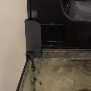 May include: A black 3D-printed plastic bracket mounted on the corner of a glass aquarium tank. The bracket features a textured surface and is positioned beneath the black plastic lid of the tank, which contains water and a dark rock.