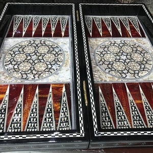 May include: A wooden backgammon board with intricate inlaid designs. The board is made of dark wood with a black and white chequered border. The playing surface is decorated with a circular pattern of mother-of-pearl and wood.