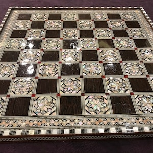Puede incluir: Un tablero de ajedrez de madera con una incrustación decorativa de nácar y acentos rojos. El tablero tiene un acabado marrón oscuro y una superficie brillante.