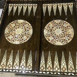 Puede incluir: Un tablero de backgammon de madera con intrincados diseños de nácar incrustados. El tablero está dividido en dos mitades, cada una con un diseño circular en el centro. El tablero es marrón con un borde marrón oscuro.