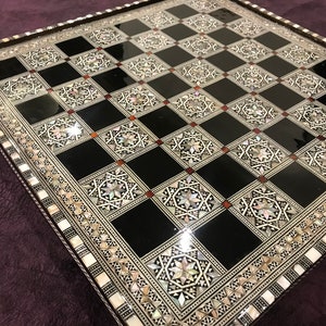 Può includere: Una scacchiera bianca e nera con un bordo decorativo in madreperla intarsiato. La scacchiera è in legno e ha una finitura lucida.