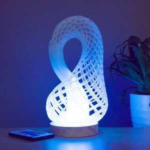 Op de afbeelding: Witte 3D-geprinte lamp met een geometrisch ontwerp, gevormd als een oneindigheidssymbool. De lamp is verlicht met blauw licht en staat op een houten tafel.