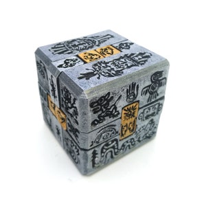 May include: A silver cube puzzle with intricate black and gold designs. The cube is made of multiple interlocking pieces and features various symbols and patterns.