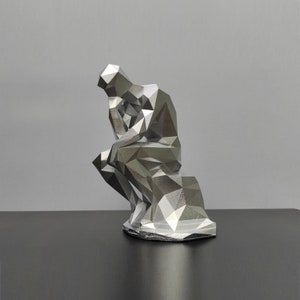 Puede incluir: Una escultura de color plateado de una persona sentada con la cabeza en la mano, en una pose de pensamiento. La escultura está hecha de muchos triángulos pequeños, lo que le da un aspecto geométrico.