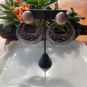 May include: A pair of silver hoop earrings with a blue and white patterned design. The earrings are on a black stand.
